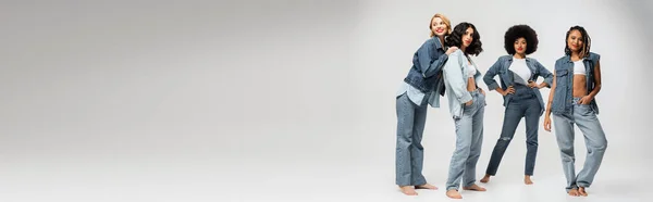 Fashionable multiracial girlfriends in blue denim wear standing barefoot on grey, horizontal  banner — Stock Photo