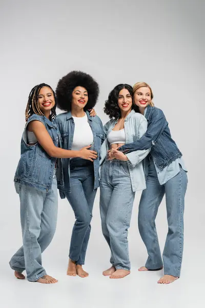 Smiley and barefoot multiethnic women in blue denim attire embracing on grey, diverse friendship — Stock Photo