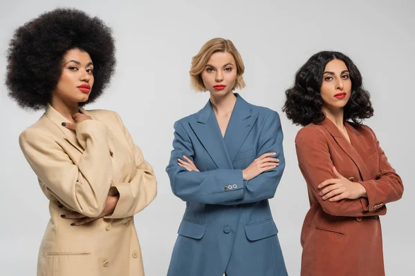 Three confident multiracial girlfriends in multicolored suits standing with folded arms on grey — Stock Photo