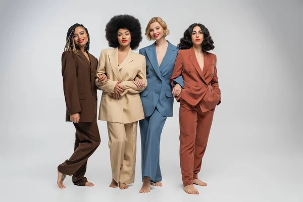Multicultural barefoot female models standing in colorful suits on grey backdrop, full length — Stock Photo