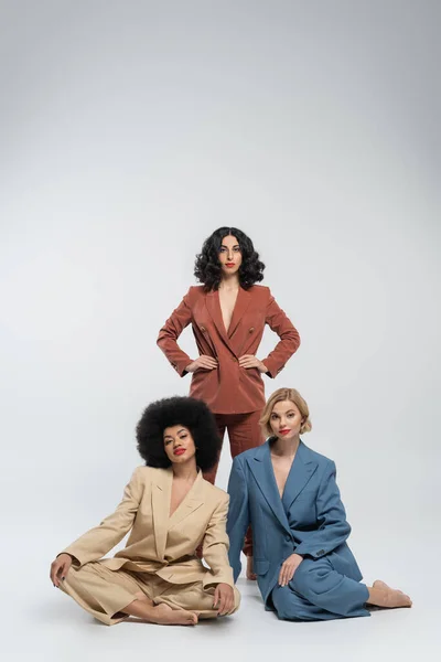 Stylish multiracial woman with hands on hips near girlfriends in colorful suits sitting on grey — Stock Photo