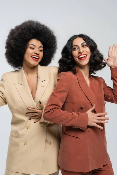 Cheerful multiracial woman waving hand near african american girlfriend on grey, colorful suits — Stock Photo