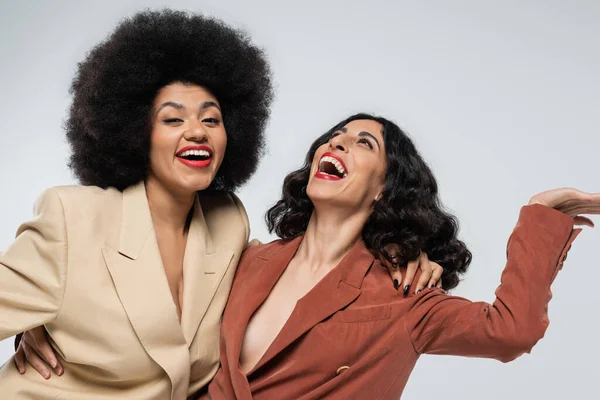 Excited multiracial woman laughing near african american girlfriend on grey, multicolored suits — Stock Photo