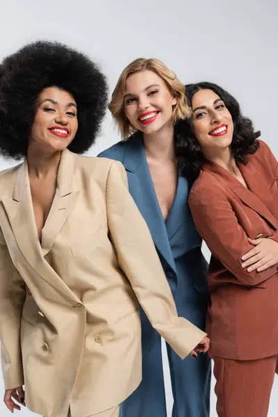 Smiley multiethnic girlfriends in multicolored suits looking at camera on grey, diverse beauty — Stock Photo