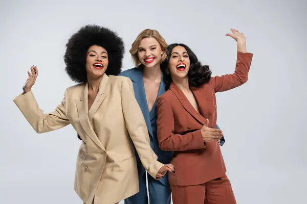 Joyeuses petites amies multiethniques en costumes colorés branchés agitant les mains à la caméra sur gris, style moderne — Photo de stock