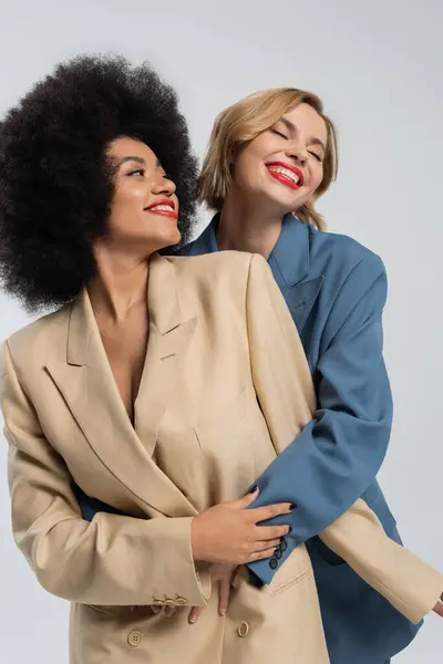 Delighted woman embracing african american girlfriend while posing in colorful suits on grey — Stock Photo
