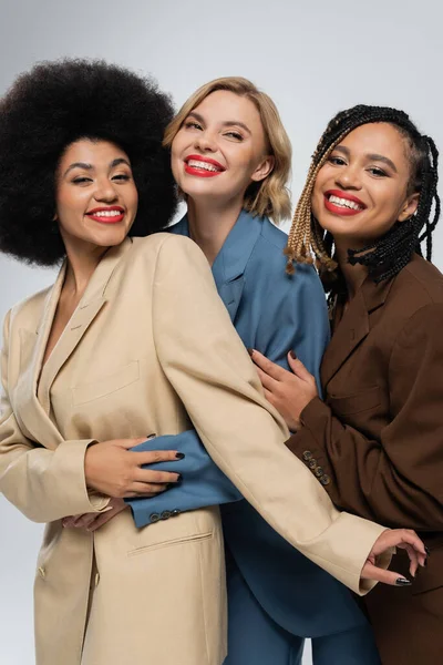 Happy multiracial girlfriends in multicolored suits embracing on grey, togetherness and style — Stock Photo