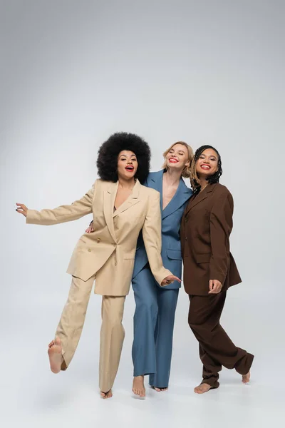 Excited barefoot multiethnic female friends in colorful suits posing barefoot on grey, full length — Stock Photo