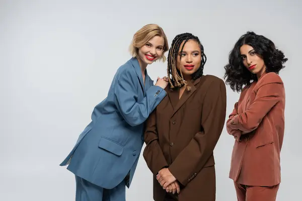 Blonde woman smiling at camera near multiethnic girlfriends in colorful suits on grey, modern style — Stock Photo