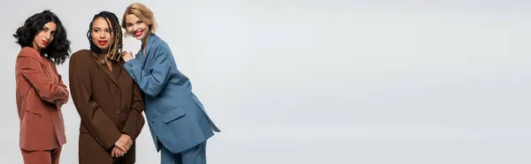 Young blonde woman smiling at camera near multiracial girlfriends in colorful suits on grey, banner — Stock Photo