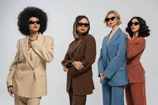 Group of trendy multiracial girlfriends in dark sunglasses and multicolored suits on grey, fashion — Stock Photo