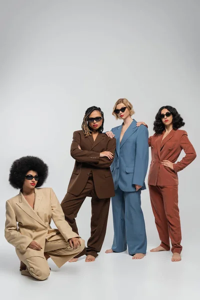 Modelos de moda multiétnica descalzo en gafas de sol oscuras y trajes de colores en gris, longitud completa - foto de stock