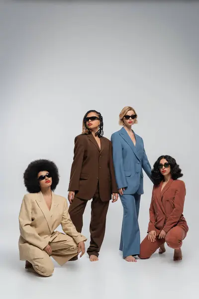 Full length of fashionable multiracial women in colorful suits and dark sunglasses on grey backdrop — Stock Photo
