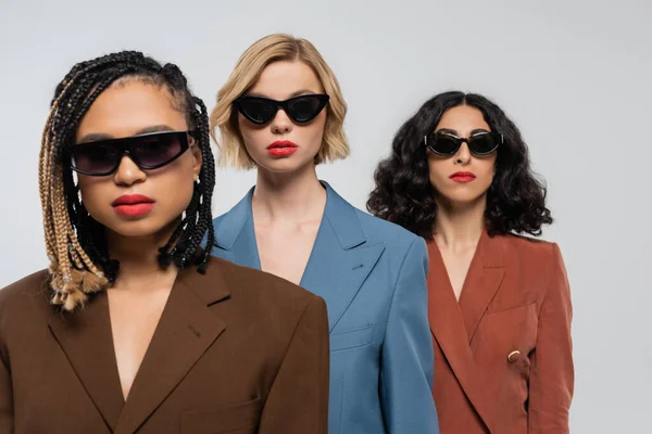 Group portrait of trendy multiracial girlfriends in dark sunglasses and colorful suits on grey — Stock Photo