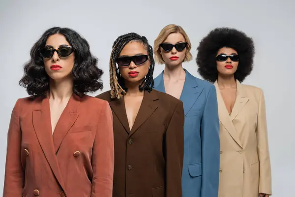 Groupe diversifié de copines multiraciales à la mode dans des lunettes de soleil sombres et des costumes colorés sur gris — Photo de stock