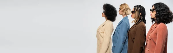 Bannière de mode, vue de côté des femmes multiethniques élégantes dans des lunettes de soleil et des costumes colorés sur gris — Photo de stock
