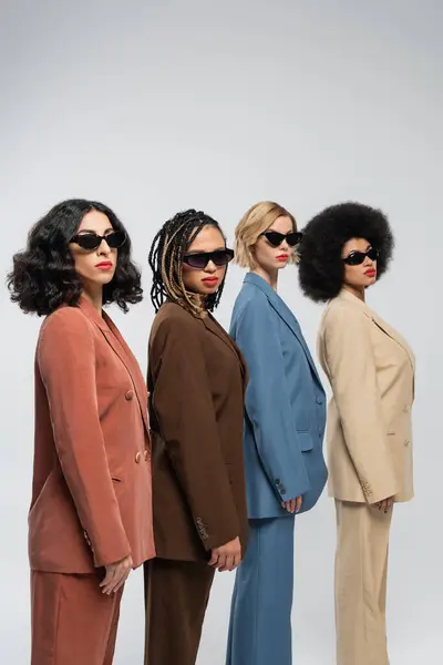 Beauty in diversity, multiracial women in sunglasses and colorful suits looking at camera on grey — Stock Photo