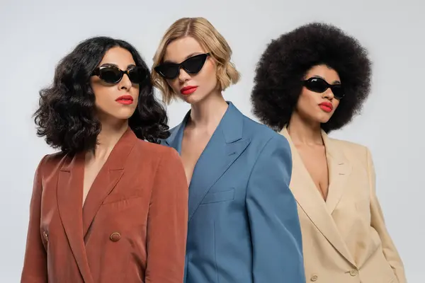 Три модні багаторасові подружки в різнокольорових костюмах і темні сонцезахисні окуляри на сірому — стокове фото