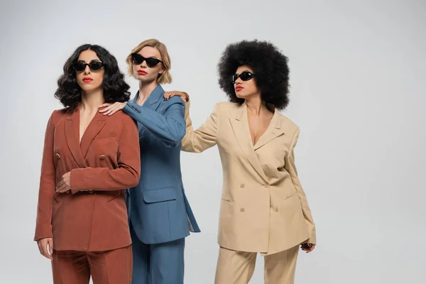 Multiethnic girlfriends in colorful suits and sunglasses touching shoulders of each other on grey — Stock Photo
