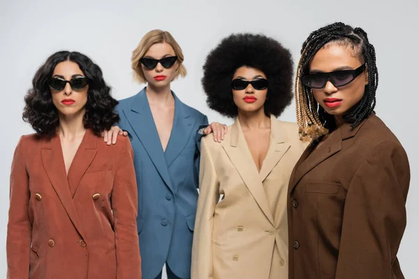 Group of trendy multiracial female friends in dark sunglasses and colorful suits on grey backdrop — Stock Photo