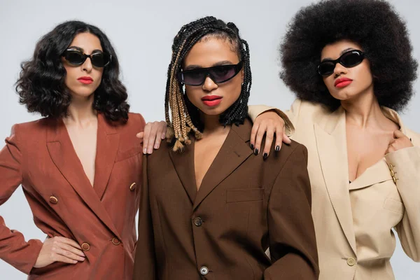 Femmes multiraciales en lunettes de soleil et costumes touchant les épaules de la petite amie afro-américaine sur gris — Photo de stock