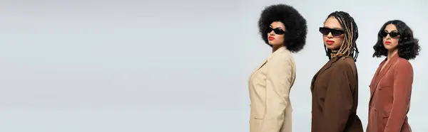 Multiracial girlfriends in trendy suits and sunglasses looking at camera on grey, fashion banner — Stock Photo