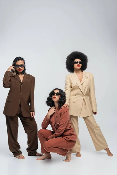 Barfuß multirassisch weibliches Modell in trendigen bunten Anzügen und dunkler Sonnenbrille auf grau, in voller Länge — Stockfoto