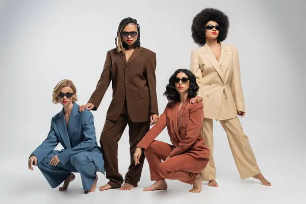 Diverse group of barefoot and stylish multiethnic girlfriends in suits and dark sunglasses on grey — Stock Photo