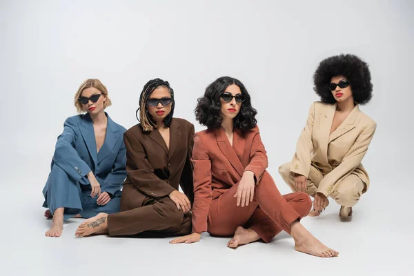 Diverse group of stylish multiracial women in dark sunglasses and colorful suits sitting on grey — Stock Photo