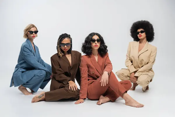 Diverse fashion, multiracial girlfriends in dark sunglasses and multicolored suits sitting on grey — Stock Photo