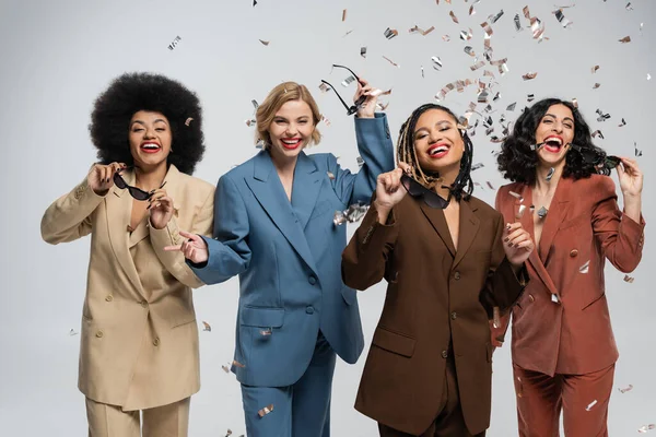 Trendy multiracial girlfriends in colorful suits rejoicing under falling confetti, party time — Stock Photo
