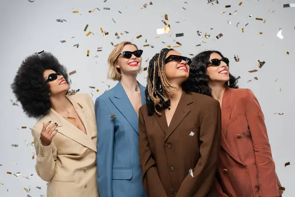 Fröhliche multirassische Freundinnen in dunklen Sonnenbrillen und bunten Anzügen, die unter festlichem Konfetti lächeln — Stockfoto