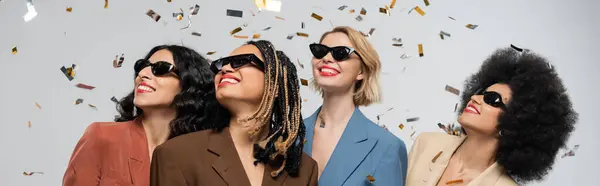Excited multiethnic girlfriends in sunglasses and colorful suits under sparkling confetti, banner — Stock Photo