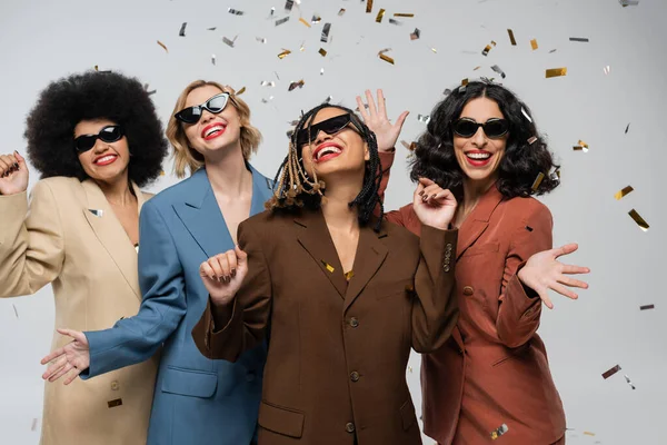 Overjoyed multiracial girlfriends in sunglasses and trendy suits under colorful confetti on grey — Stock Photo