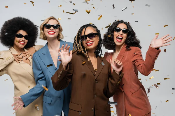 Tempo de festa, namoradas multirraciais alegres em óculos de sol e ternos sob confete festivo em cinza — Fotografia de Stock