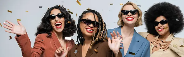 Модні багаторасові подружки в сонцезахисних окулярах і костюмах, радіючи під конфетті на сірому, банер — стокове фото