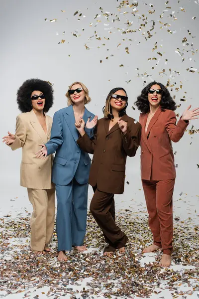 Barefoot and cheerful multiethnic models in dark sunglasses and stylish suits near confetti on grey — Stock Photo