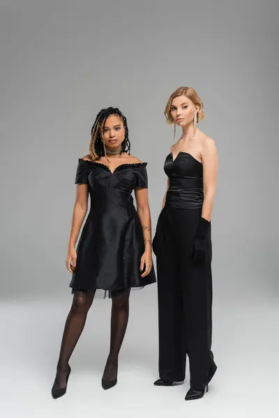 Full length of young multiracial women in black elegant attire on grey backdrop, diverse beauty — Stock Photo