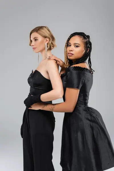 Elegant african american woman in black attire embracing waist of blonde glamour friend on grey — Stock Photo