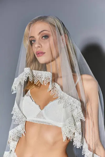 Portrait of attractive young woman in white lingerie with veil looking at camera on gray backdrop — Stock Photo