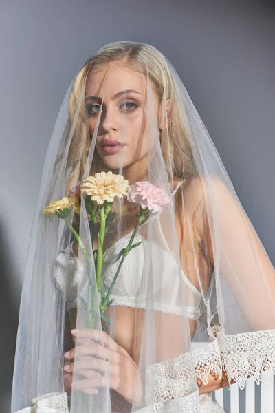 Vertical shot of attractive young woman wearing veil posing with flowers in hands looking ta camera — Stock Photo