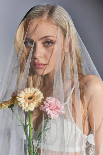 Vertical shot of sexy woman with blonde hair in white veil holding flowers and looking at camera — Stock Photo