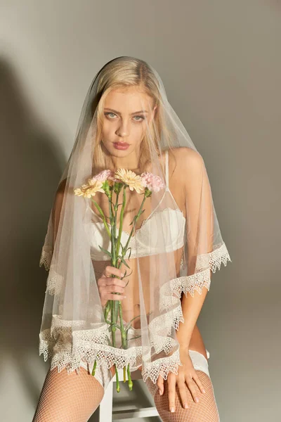Vertical shot of sexy young woman in erotic lingerie with veil holding flowers sitting on tall chair — Stock Photo