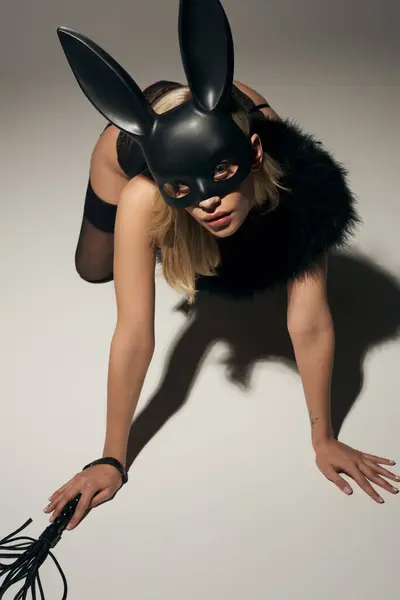 Atractiva mujer joven en sexy máscara de conejo negro posando seductor en el suelo y mirando a la cámara - foto de stock