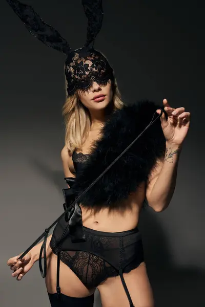Tiro vertical de mujer joven sexy en lencería negra con piel sintética y máscara de conejo con látigo en las manos - foto de stock