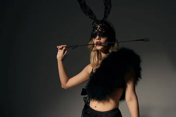 Сексуальная молодая женщина в привлекательном черном наряде с маской кролика, позирующая и держащая в зубах кнут — стоковое фото