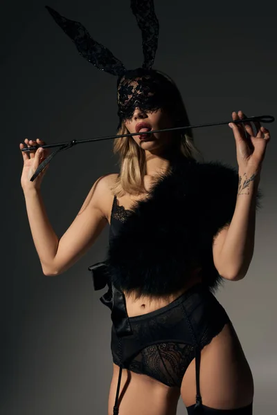 Atractiva mujer rubia en máscara de conejo y lencería de encaje negro tocando látigo con su lengua - foto de stock
