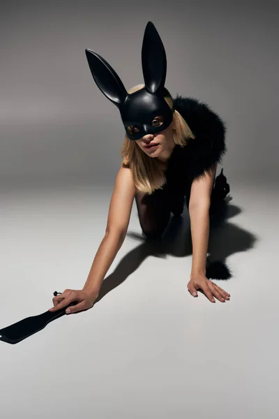 Attraente donna in maschera di coniglio nero e lingerie di pizzo in piedi sulle ginocchia sul pavimento con bdsm frusta — Foto stock