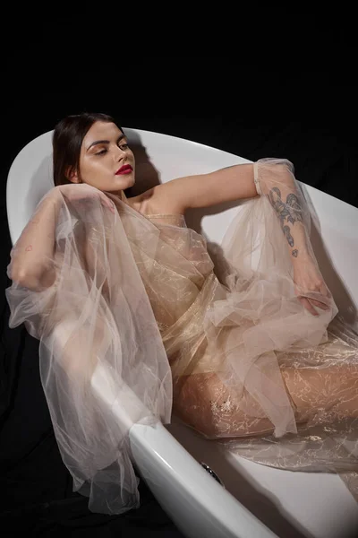High angle view of graceful young model in transparent dress while lying on bathtub on black — Stock Photo