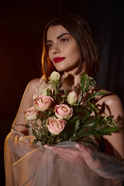 Charming young woman in romantic transparent dress holding blooming roses on black backdrop — Stock Photo